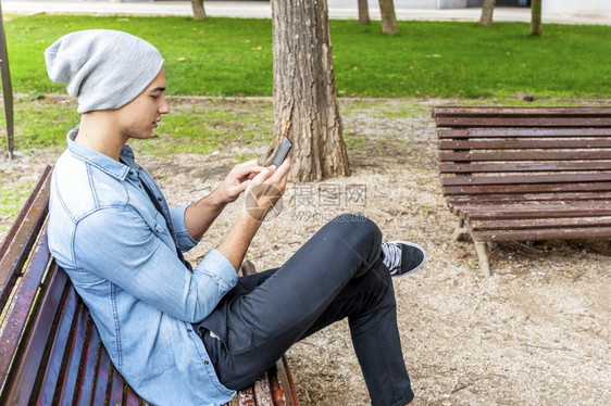 快乐的青年男子在户外闲坐时穿着手持机浏览忙碌的时髦图片