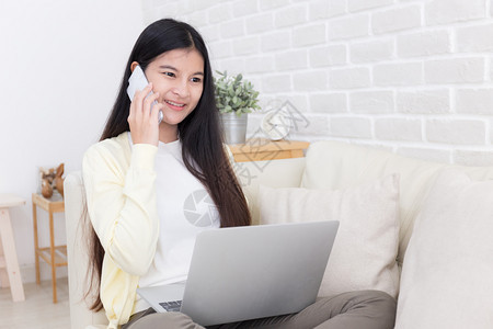 在白色房间沙发上使用智能手机和笔记本电脑的亚洲妇女在线泰国学生图片