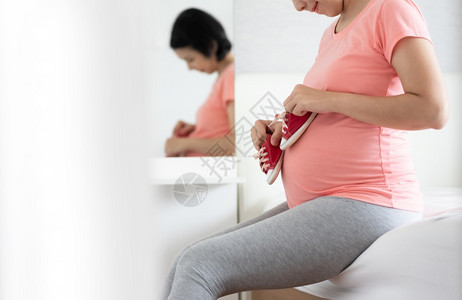 女士期待肚子穿着粉红衬衫健康孕妇在卧室怀期间坐在床上休息放松时微笑地将小婴儿红鞋抱在贝利亚洲快乐母亲身上图片