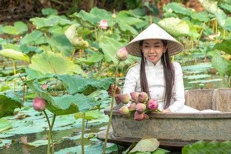 身穿白色传统越南女美丽亚西雅人身着AoWai和越南农民帽子穿着白传统越南服装坐在花莲湖中的木船上手摘莲花自然木制的脸图片