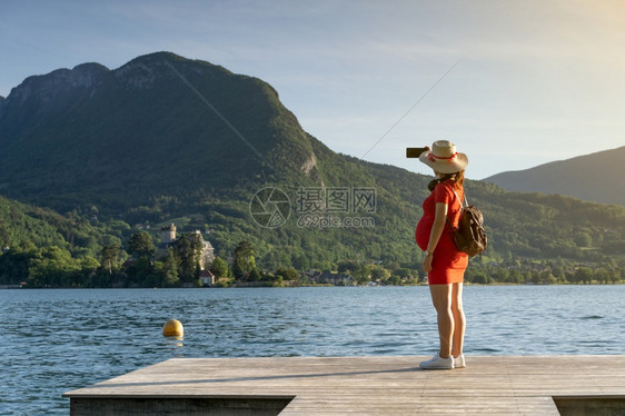 在大山前的码头上湖边自拍穿着跟踪帽子和红礼服的女孩在大山前面的码头上夏天坐着景观图片
