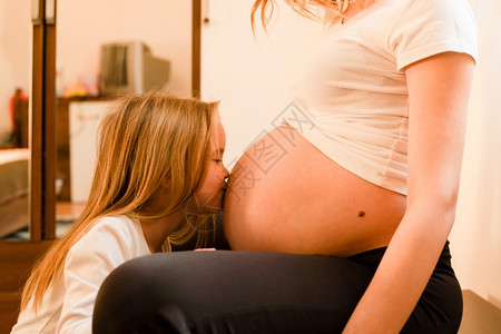 女儿在家中站立亲吻孕妇肚子等待婴儿的到来女孩在家小儿站着亲吻母肚子怀孕等待婴儿的到来未出生妈拥抱图片