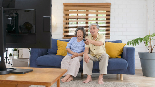 在家中客厅看电视时持遥控器的老年亚裔夫妇在有幸福老年人退休和生活正常的安居室观看电视亚洲坐着老化图片