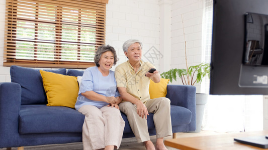 在家中客厅看电视时持遥控器的老年亚裔夫妇在有幸福老年人退休和生活正常的安居室观看电视男人闲暇沙发图片