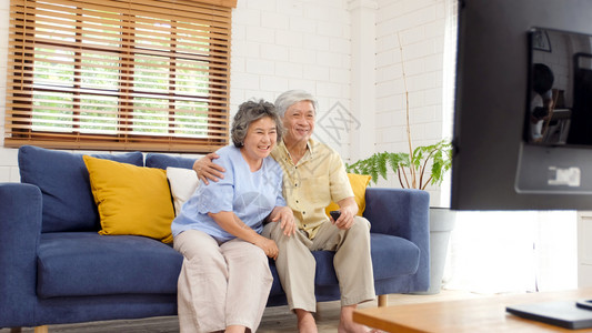 人们在家中客厅看电视时持遥控器的老年亚裔夫妇在有幸福老年人退休和生活正常的安居室观看电视妻子乐趣图片