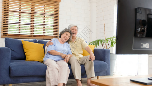 在家中客厅看电视时持遥控器的老年亚裔夫妇在有幸福老年人退休和生活正常的安居室观看电视陈年60代控制图片