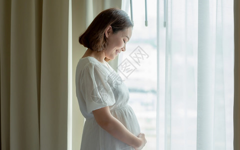 幸福女士身着白裙子站在卧室窗户旁边的亚洲美丽孕妇母亲婴儿和怀孕概念组织妇女婴儿和怀孕概念分娩图片