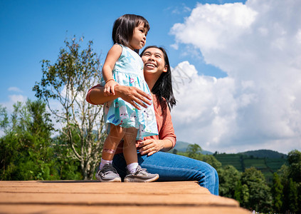 快乐的母亲与女儿坐在木地板上树有山地背景在夏日蓝天下色的裙子成年人图片