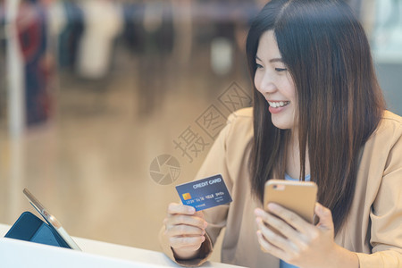 无卡支付亚洲妇女利用信卡和移动电话在百货商店通过服装背景技术钱财包和网上支付概念信用卡模型在线购物亚洲妇女使用信卡和移动电话在服装店商进背景