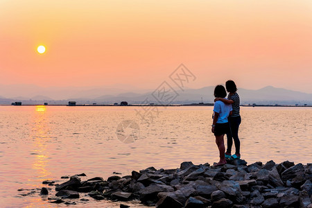 一种山看着水边美丽的自然景观湖边日落时阳光照在明亮的橙色天空中泰国KrasiaoDamSuphanBuri母亲和女儿观看海边的日图片