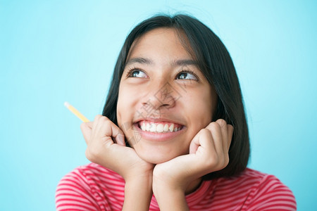 手工作室接触亚裔女孩在背景蓝色孤立地边上思考和微笑亚洲儿童快乐地握着铅笔摸下巴一边看图片