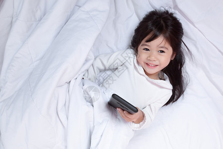 卧室亚洲小孩玩智能手机得开心早上躺在床坐软枕头笑起来感觉很快乐的喜悦幸福说谎图片