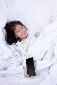 微笑亚洲小孩玩智能手机得开心早上躺在床坐软枕头笑起来感觉很快乐的喜悦应用程序网络图片