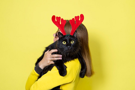 猫女孩猫咪穿黄色裙子的女孩拿着她心爱的毛茸滑稽黑猫戴着圣诞驯鹿角头带戴着黄色背景的圣诞猫黄色背景脸节背景
