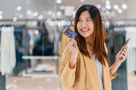亚洲妇女利用信卡和移动电话在百货商店通过服装背景技术钱财包和网上支付概念信用卡模型在线购物亚洲妇女使用信卡和移动电话在服装店商进图片