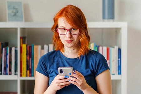 抓住自然乐趣穿着眼镜的模糊书架面前拿着移动智能手机的蓝色T恤衫和橙头发与蓝T恤在家中面对着模糊的书架门前举起行动智能电话图片