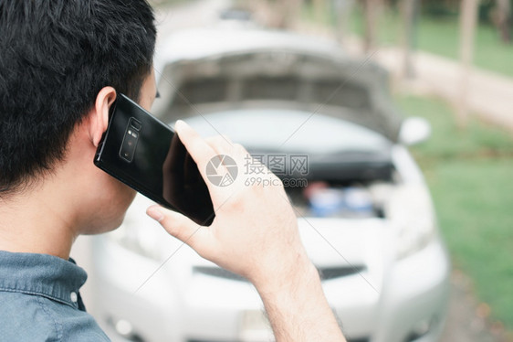 街道警告司机使用移动电话的亚洲男子在街头汽车故障后要求援助车辆发动机问题概念或事故以及专业技工的紧急帮助在交通失灵之后使用移动电图片