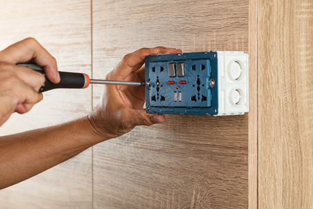 电工正在用螺丝起子把电源插到木墙上的塑料盒职业电气检查图片