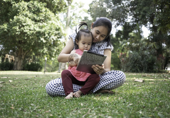 亚洲母亲和女儿坐在花园的草地上欣喜看着智能手机并坐在花园的草地上家庭孩子等待图片