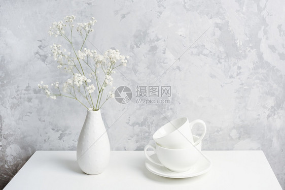 老的早晨结石花瓶中的一束小满天星和灰色混凝土墙上的桌子两个白色茶杯或咖啡复制空间前视图图片