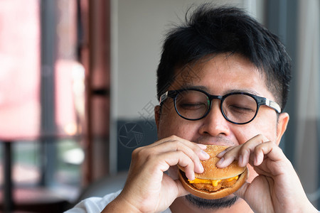 亚洲男人在快餐厅吃汉堡包享受着美味的食物穿着白色T恤和眼镜的人拿着汉堡包享受的口味笑成人桌子莴苣图片