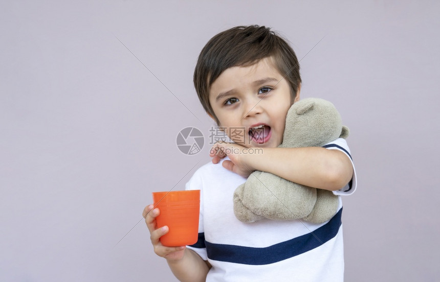 手关心保持长片肖像可爱的小男孩喝橙子汁从塑料杯的橙汁在浅紫背景的轻玻璃上健康的45岁男孩喝着混合果汁和泰迪熊图片