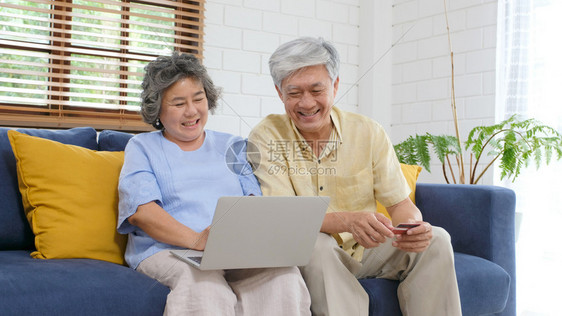 通过使用膝上型计算机和家庭客厅信用卡退休者技术生活方式等在家中用笔记本电脑和信用卡在线购物的老年亚裔夫妇放松女士图片