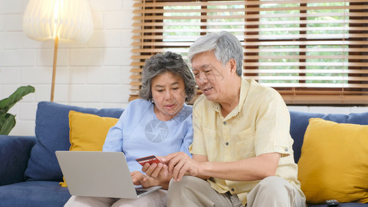 爱长椅通过使用膝上型计算机和家庭客厅信用卡退休者技术生活方式等在家中用笔记本电脑和信用卡在线购物的老年亚裔夫妇女士图片