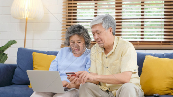通过使用膝上型计算机和家庭客厅信用卡退休者技术生活方式等在家中用笔记本电脑和信用卡在线购物的老年亚裔夫妇洲人支付电子的图片
