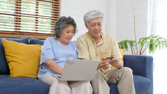 保持买成人通过使用膝上型计算机和家庭客厅信用卡退休者技术生活方式等在家中用笔记本电脑和信用卡在线购物的老年亚裔夫妇图片