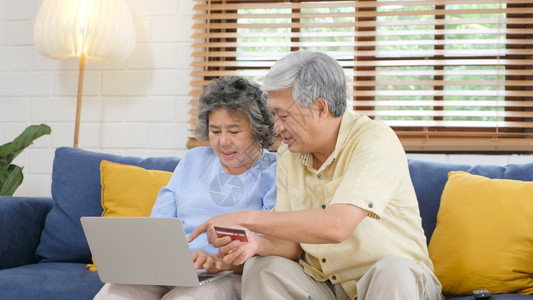 经过爱亚洲人通使用膝上型计算机和家庭客厅信用卡退休者技术生活方式等在家中用笔记本电脑和信用卡在线购物的老年亚裔夫妇图片