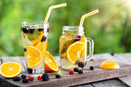 杯子和罐加橘鸡尾柠檬水浆果放在木制桌边阳光明媚的白天玻璃还有橙子鸡尾柠檬水和浆果甜的鸡尾酒多汁图片