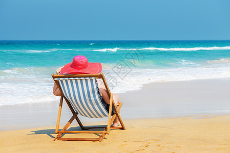 坐在甲板椅上看着海景和旅行概念快乐的女士在沙滩上穿着红色太阳浴衣坐在甲板椅上海度假帽子女图片