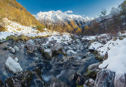 树木绿色阳光山河在岩石之间流动挪威秋天的快速水金树雪覆盖的山脉图片