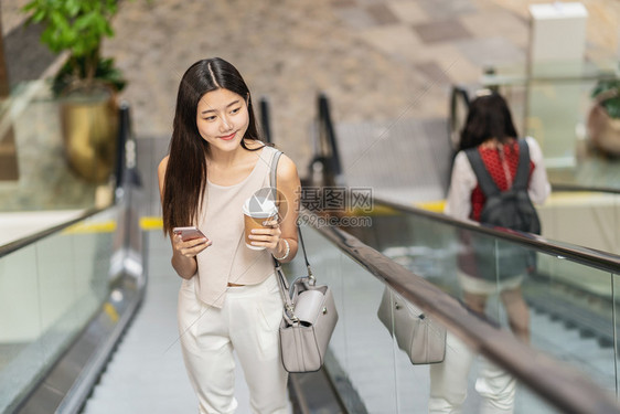 人群使用智能移动电话和在百货商店步行扶梯的亚洲年轻女乘客在城市japanesechinese朝韩生活方式和日常commuter及图片