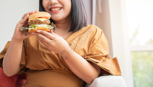 快速地身体狂欢饥饿的超重妇女笑着拿汉堡包坐在客厅里她非常快乐并享受吃餐狂食饮紊乱概念图片