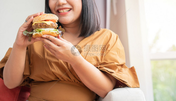 快速地身体狂欢饥饿的超重妇女笑着拿汉堡包坐在客厅里她非常快乐并享受吃餐狂食饮紊乱概念图片