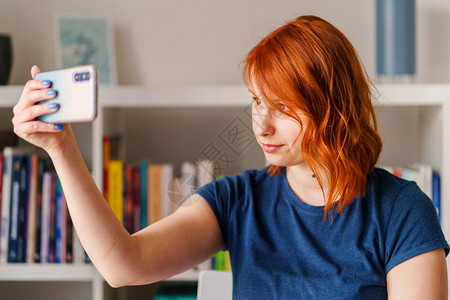 明亮的快乐大学在家清晨时书架橙色头发前或工作使用手机智能拍摄蓝色T恤自照片的美人脸部女caucasiansian女孩肖像图片