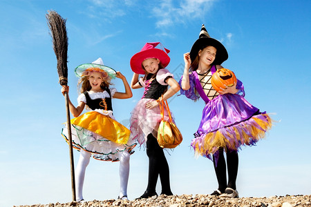 女孩们露齿三个美丽的微笑小女孩长发在彩色服装中看着摄影机与扫帚和捣弄或招待袋人们图片