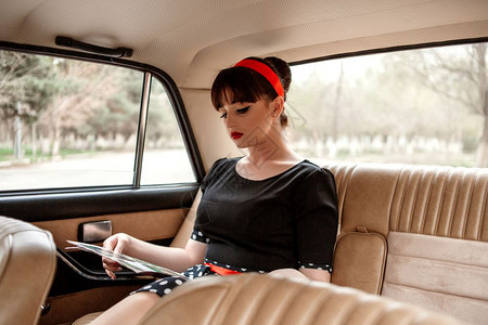 车辆女白种人身着黑色古年礼服的高加索轻美女肖像在旧车沙龙上装扮阅读一本旧杂志图片
