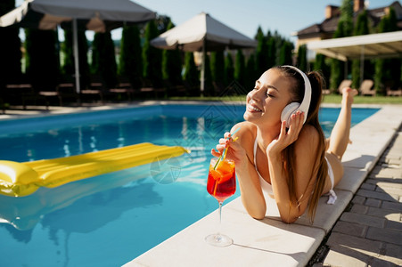 游泳的在池边缘听音乐和喝汽水的妇女耳机在游泳池边缘听到音乐和喝小鸡尾酒的人欢乐快们在暑假泳池边户外的节日派对上玩得开心妇女休闲人图片