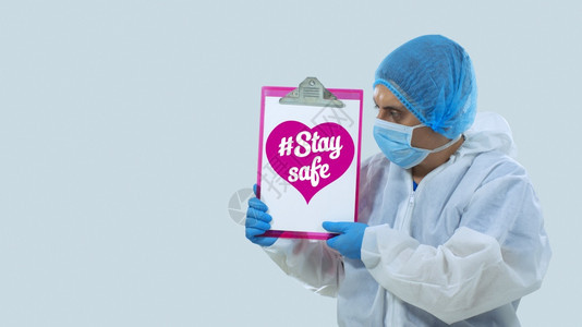 脸颜色戴面罩和蓝手套的医生拿着一个带有保持安全信息的白板在色背景上的粉红心脏内刻字图片