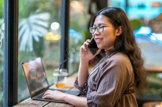 微笑淑女商人在咖啡店用笔记本电脑工作时用咖啡杯和智能手机如背景通信概念等在咖啡店里捡起智能便携机的空闲手写作抓住图片