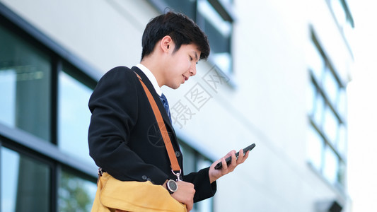 保持联网青年商人使用移动电话应程序在办公室外与背景的摩天大楼通短信年轻商人持有智能手机从事商业工作的青年男子人们图片