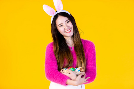穿着兔子耳朵的亚洲快乐女青年手拿着一个篮子配色彩多的复活节鸡蛋和侧眼看着摄影机在黄色空版间工作室中被孤立庆祝假期时髦图片