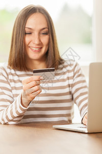 尽管在线的笔记本电脑在家网上购物时微笑着的年轻女士手里握着信用卡手持图片