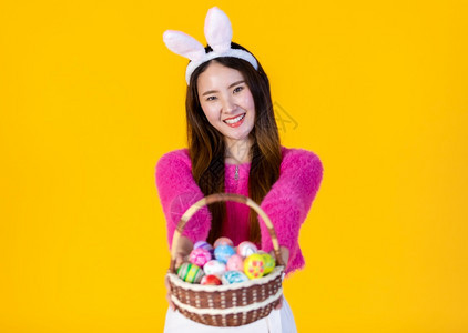 模型庆祝穿着兔子耳朵的亚洲快乐女青年手拿着一个篮子配彩色复活节鸡蛋在黄色空版间工作室背景上孤立无闻美丽的图片