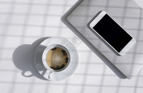 技术现代的白色陶瓷杯中草药茶表面的阳光和网格阴影图案白桌顶上有空智能手机和灰色笔记本电脑放松茶叶时间概念空白的图片