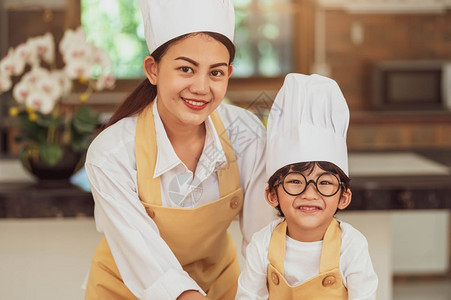 高兴地制服孩子带着眼镜的肖像可爱小亚洲男孩厨师和他母亲在家中烹饪厨房里寻找照相机人们快乐而有趣的生活方式和家庭自制食品成份概念人图片