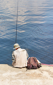淡水晴天池塘穿浅色巴拿马顶帽的渔民和穿着背心的渔民单独坐在水泥码头上阳光明日的垂直图像中水边有一根鱼杆靠水坐着一条渔棍个戴着巴拿图片